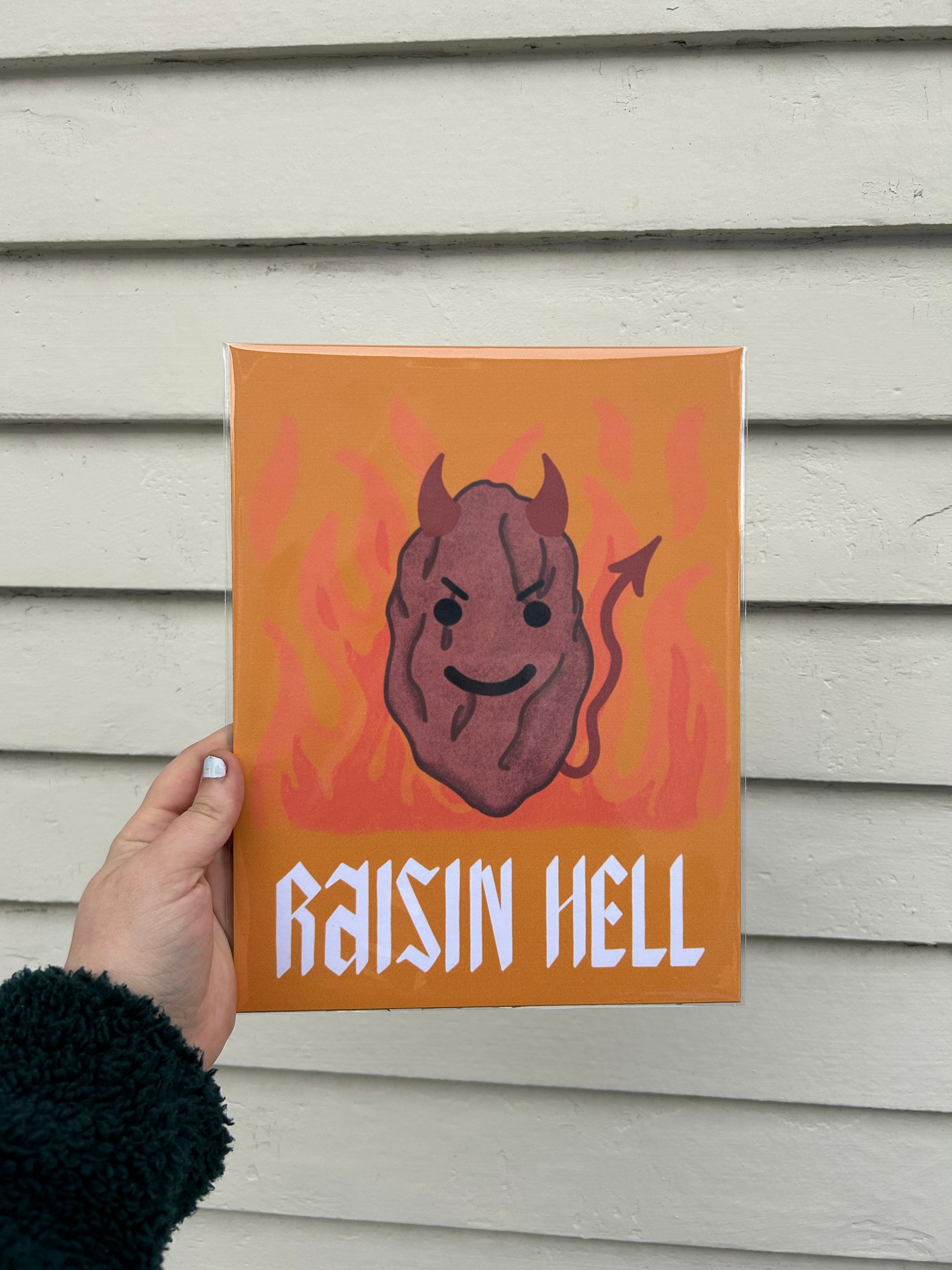 A hand holds a print enclosed in a clear plastic sleeve over a white background. The print image is as follows; on a dark yellow background, standing in front of flames, is a large raisin. The raisin is wearing devil horns and has a devil tail with a mischievous expression. Below the raisin are the words "RAISIN HELL" in white text.