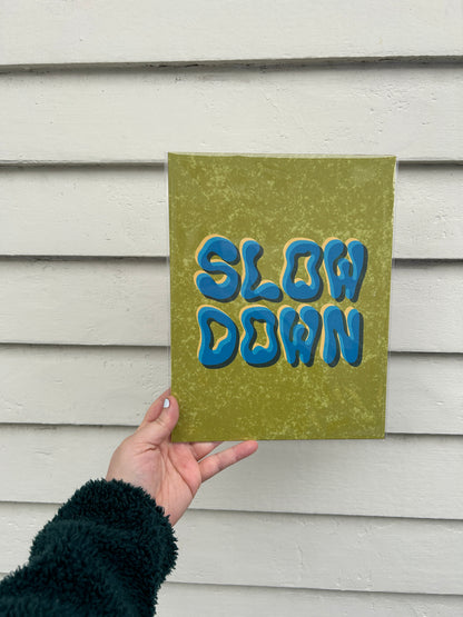 A hand holds a print enclosed in a clear plastic sleeve over a white background. The print image is as follows; on an olive green speckled background, the words "slow down" appear three times. They are layered on top of each other, methodically askew so that a small portion of the previous layer peeks through.