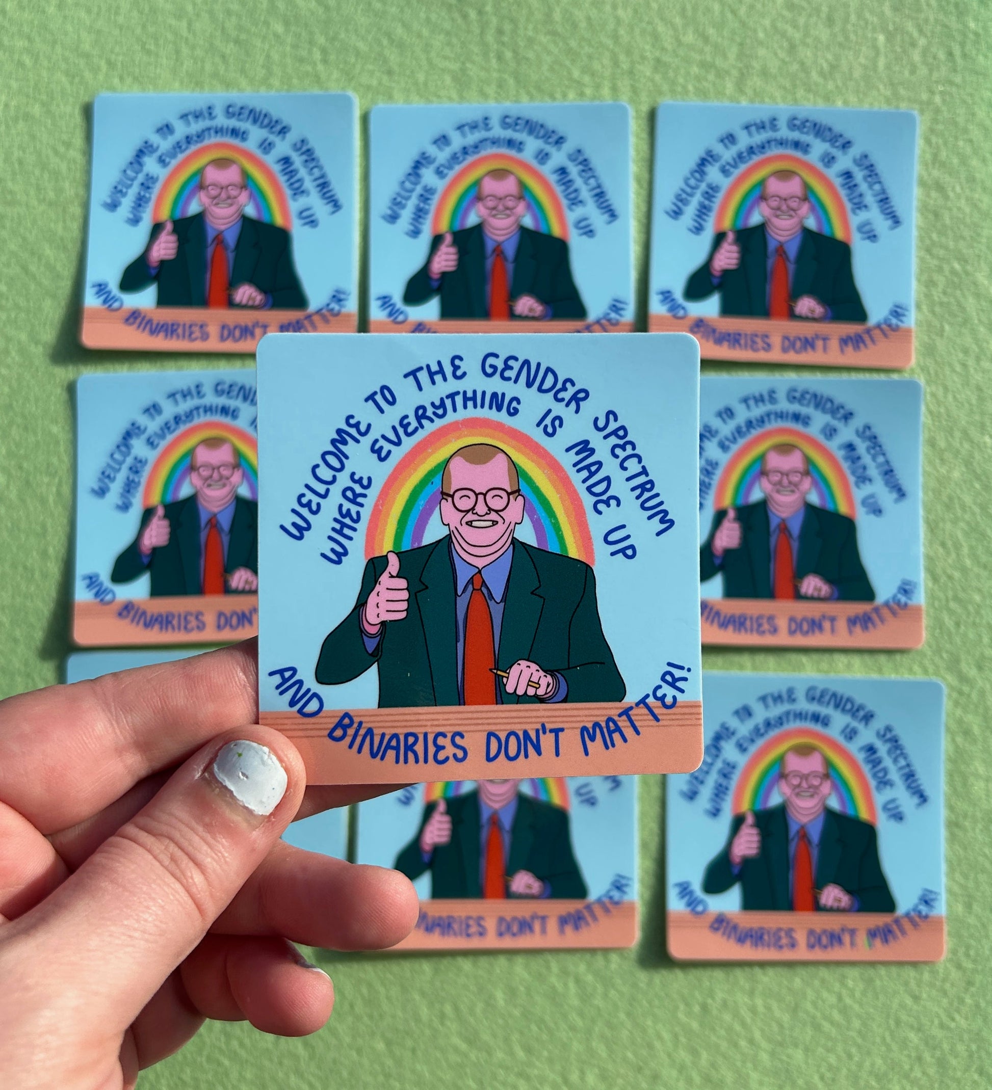 A sticker is held above a grid of other identical stickers. They all have an image of a smiling Drew Carey sitting at a desk and giving a thumbs up, with the words "Welcome to the gender spectrum, where everything is made up and binaries don't matter" above and below him.