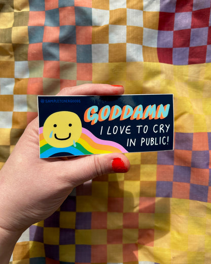 A hand holds out a sticker from the bottom left corner. The sticker reads "goddamn I love to cry in public" and has a picture of a crying smiling face above a rainbow.