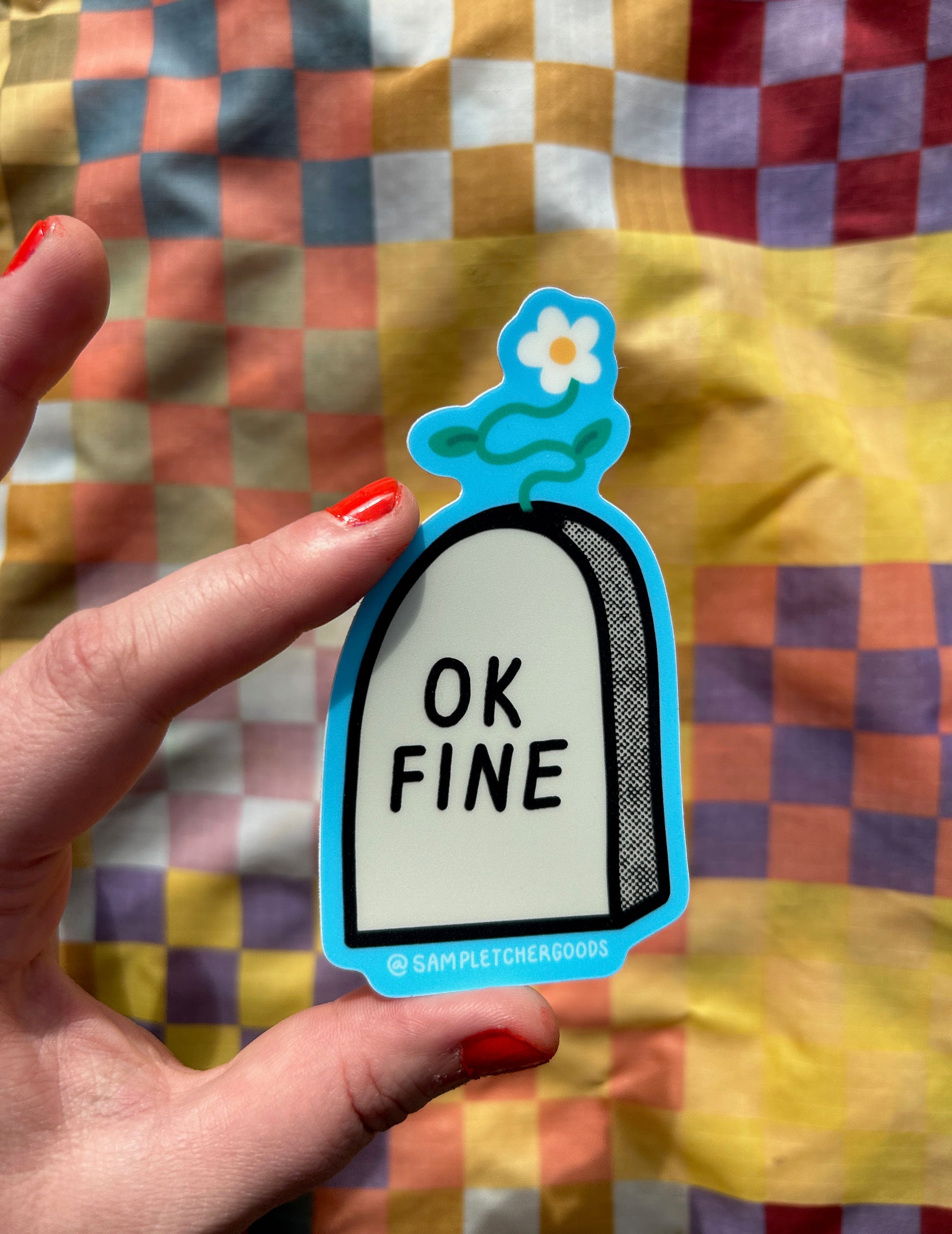 A hand holds a sticker out over a colorful background. The sticker design is an image of a gravestone with a small flower growing from the top of it. The gravestone reads "OK FINE"