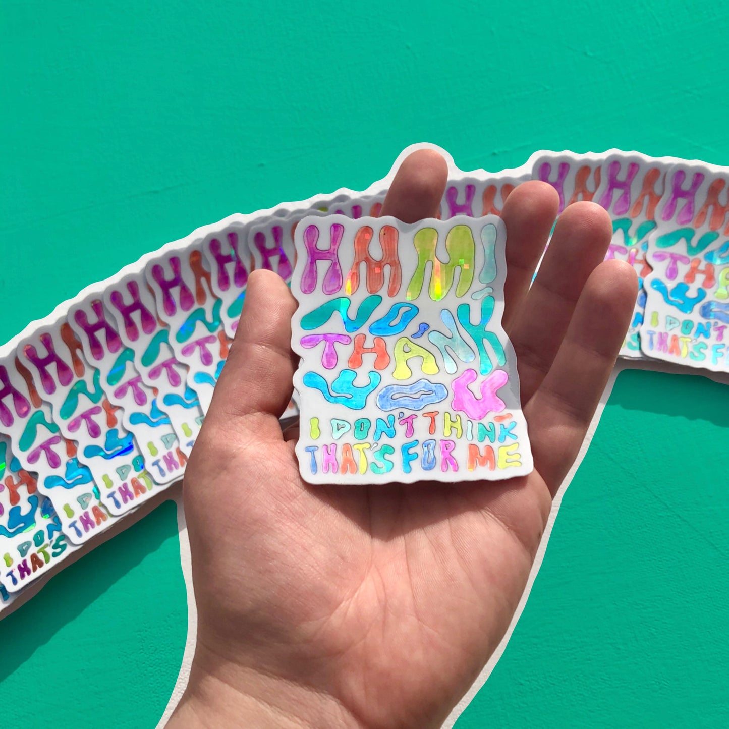 A hand holds a single sticker upon a background of identical stickers. All the stickers read "Hmm! No thank you, I don't think that's for me" in holographic rainbow font.