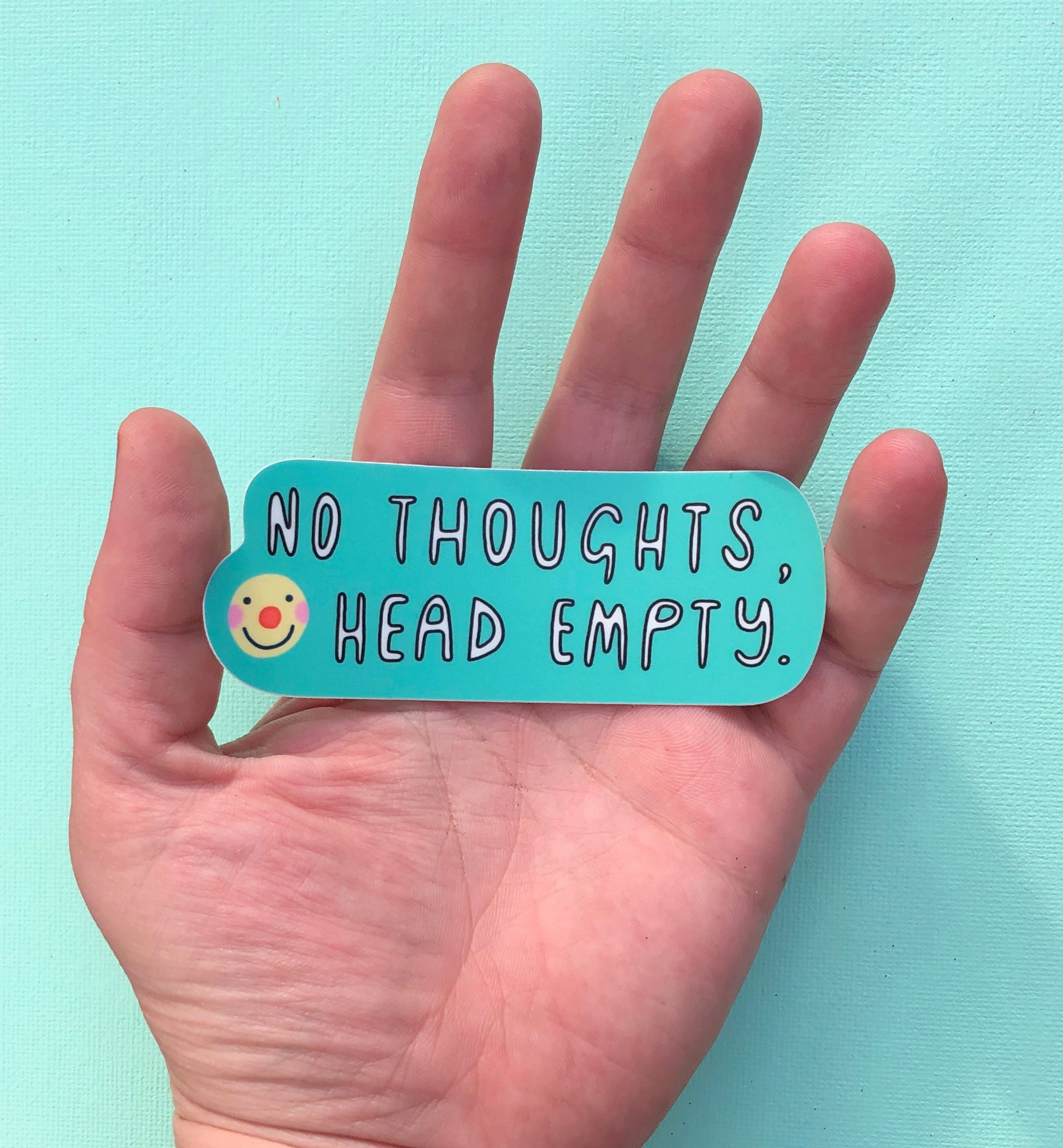 A palm facing up, with a single sticker laying on it, is in the middle of the photo. The sticker reads "no thoughts, head empty" on a mint background with an image of a smiley face wearing clown make up next to the words.