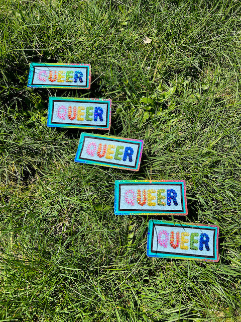 Five identical stickers placed in grass, all reading "QUEER" with the letters being made from sequins. The letters and the border have a holographic glitter sheen.