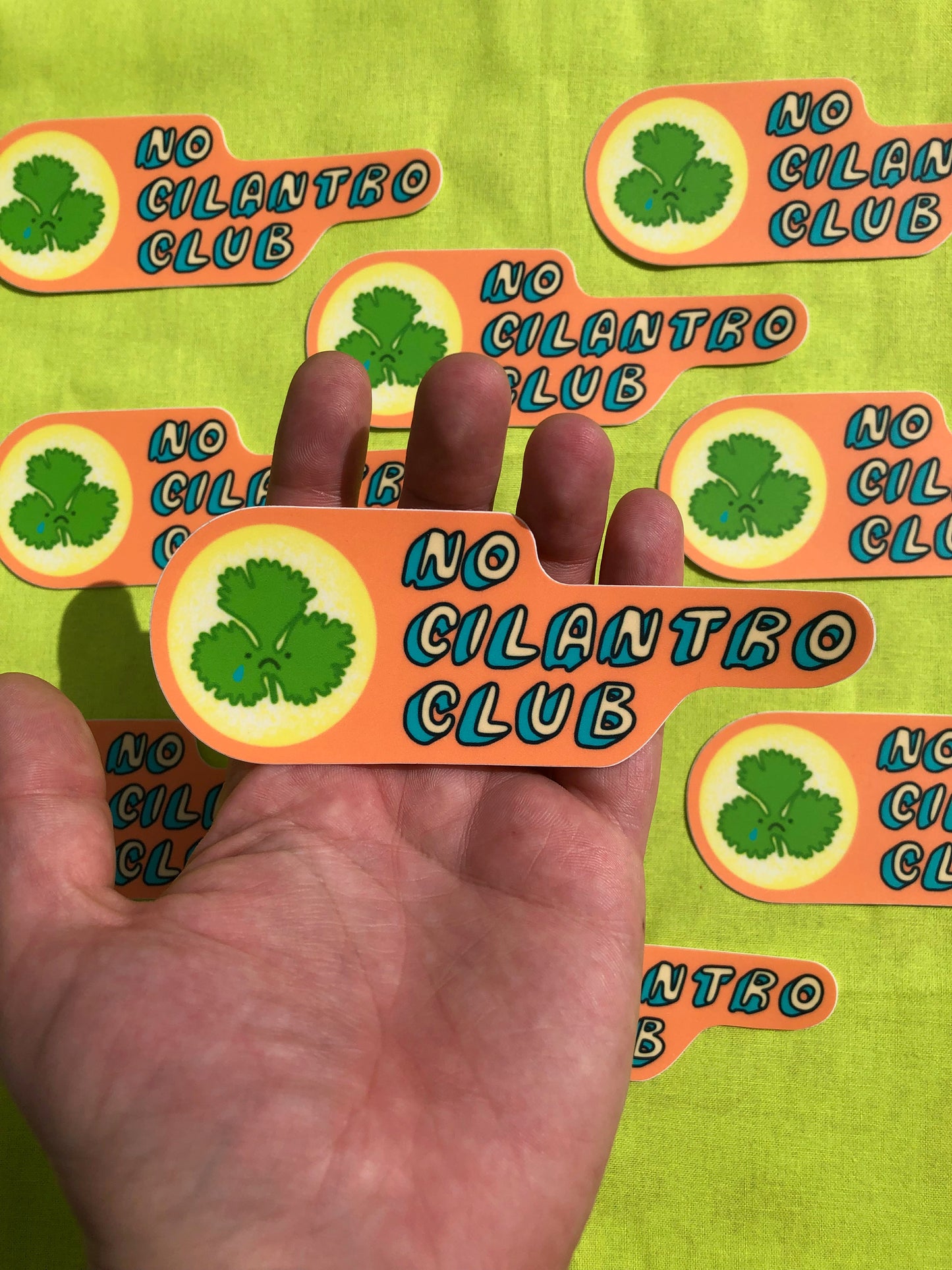 A hand holds out a single sticker above a background of many stickers. They all read "No Cilantro Club" with bubble letters on an orange background next to a picture of a cilantro leaf with a crying sad face.