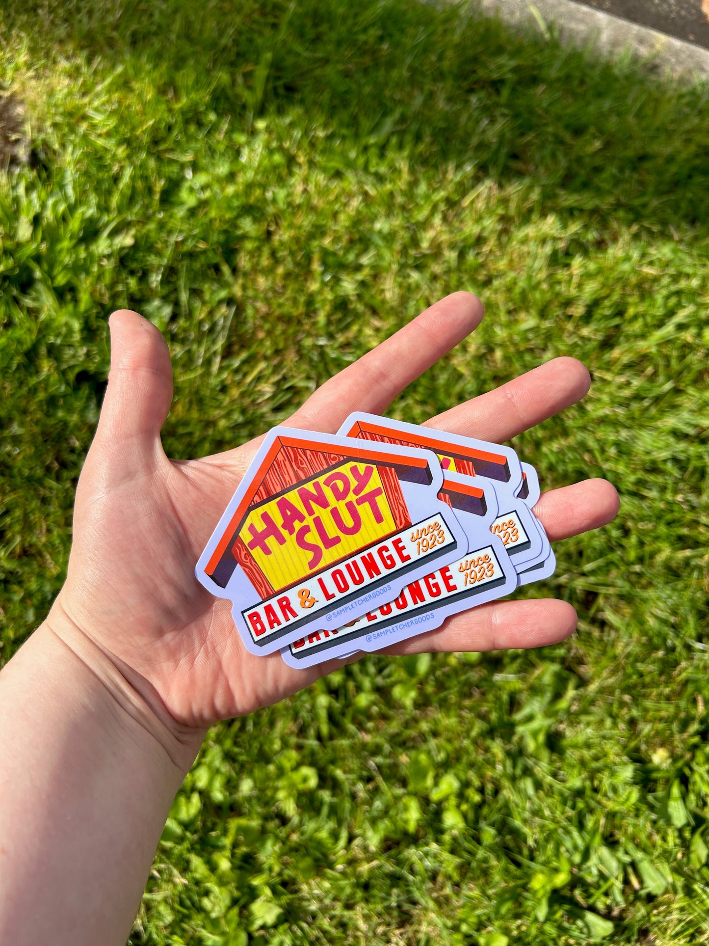 A hand holds out a pile of identical stickers above a background of grass. The sticker design is based on a local Portland, Oregon dive bar called the Sandy Hut. The sticker is an image of the bar's sign but with the first two letters swapped and an "L" added so it reads "Handy Slut, bar & lounge since 1923"