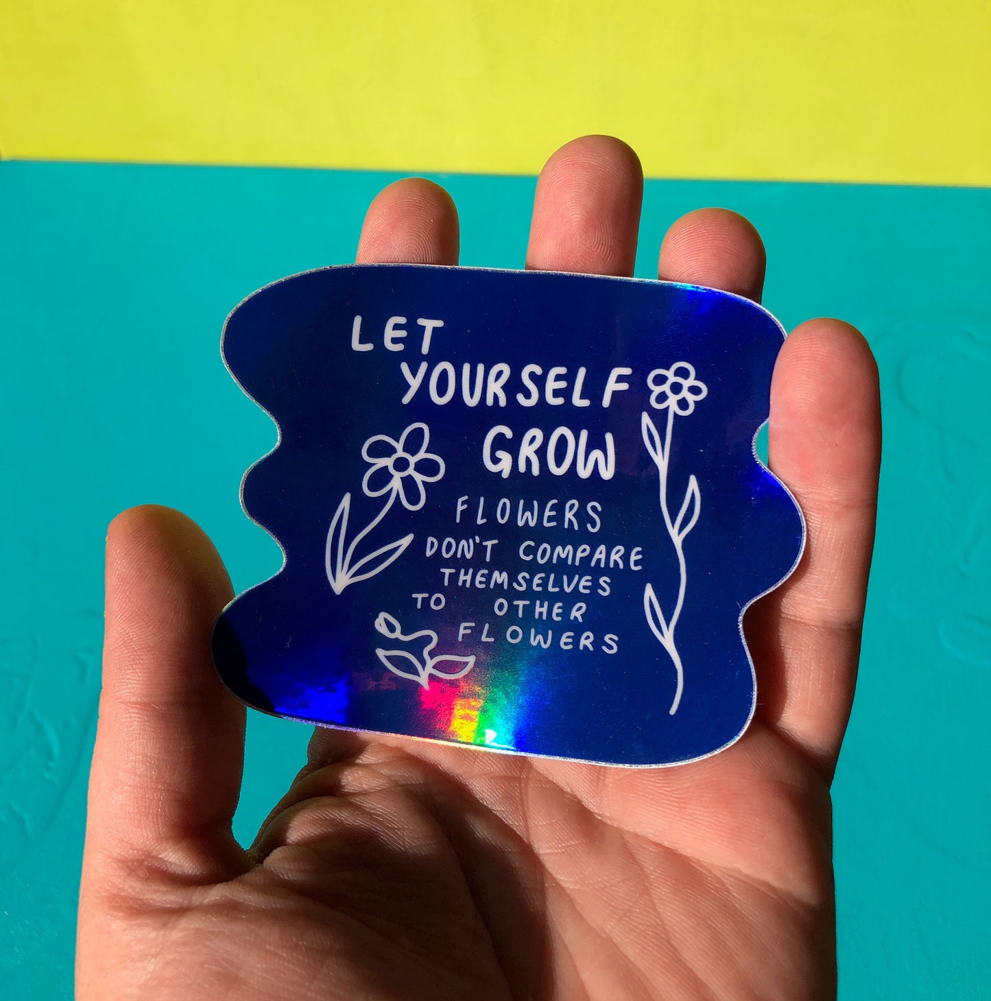 A single sticker is held in front of a teal and green background. The sticker itself is royal blue and has a vibrant holographic sheen. It reads "Let Yourself Grow; flowers don't compare themselves to other flowers" in white text with three simple images of flowers in various states of growth also in white, on both sides of the text.